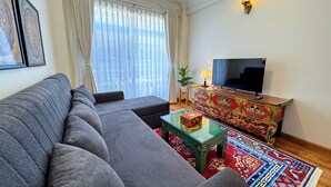 Ein Wohnzimmer mit grauem Sofa, einem Fernseher auf einer dekorativen Kommode, einem kleinen Tisch mit einer Schachtel und einem farbenfrohen Teppich.
