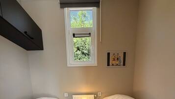 Une petite pièce bien éclairée, avec deux lits simples, un radiateur, une fenêtre donnant sur un espace verdoyant et un tableau encadré au mur.