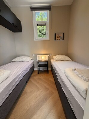Quarto pequeno e bem iluminado com duas camas individuais, um radiador, uma janela com vista para o verde e um quadro na parede.