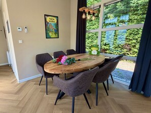 Uma sala de jantar com uma mesa de madeira redonda, seis cadeiras e vista para o verde através de grandes janelas.