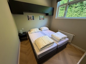 A bedroom with two beds, a nightstand, and a window with a view of greenery. - Lodge 45 | Luxe lodge met airco | Zennewijnen, Betuwe (Zennewijnen)
