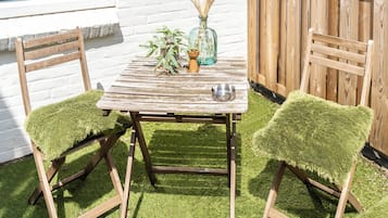 A wooden table with a rustic design, two chairs with green upholstered seats, a vase with dried plants, and a metal bowl on the table.