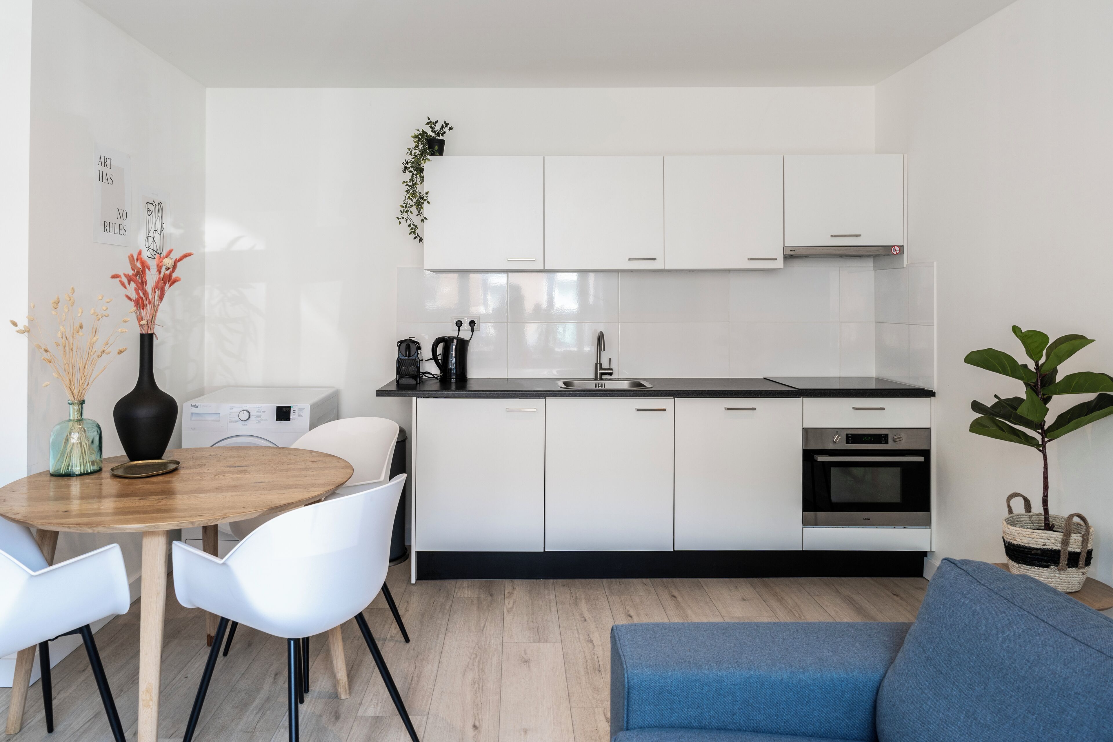 A modern kitchen with white cabinets, a black countertop, and a built-in oven. There is a dining table with white chairs and a blue sofa.