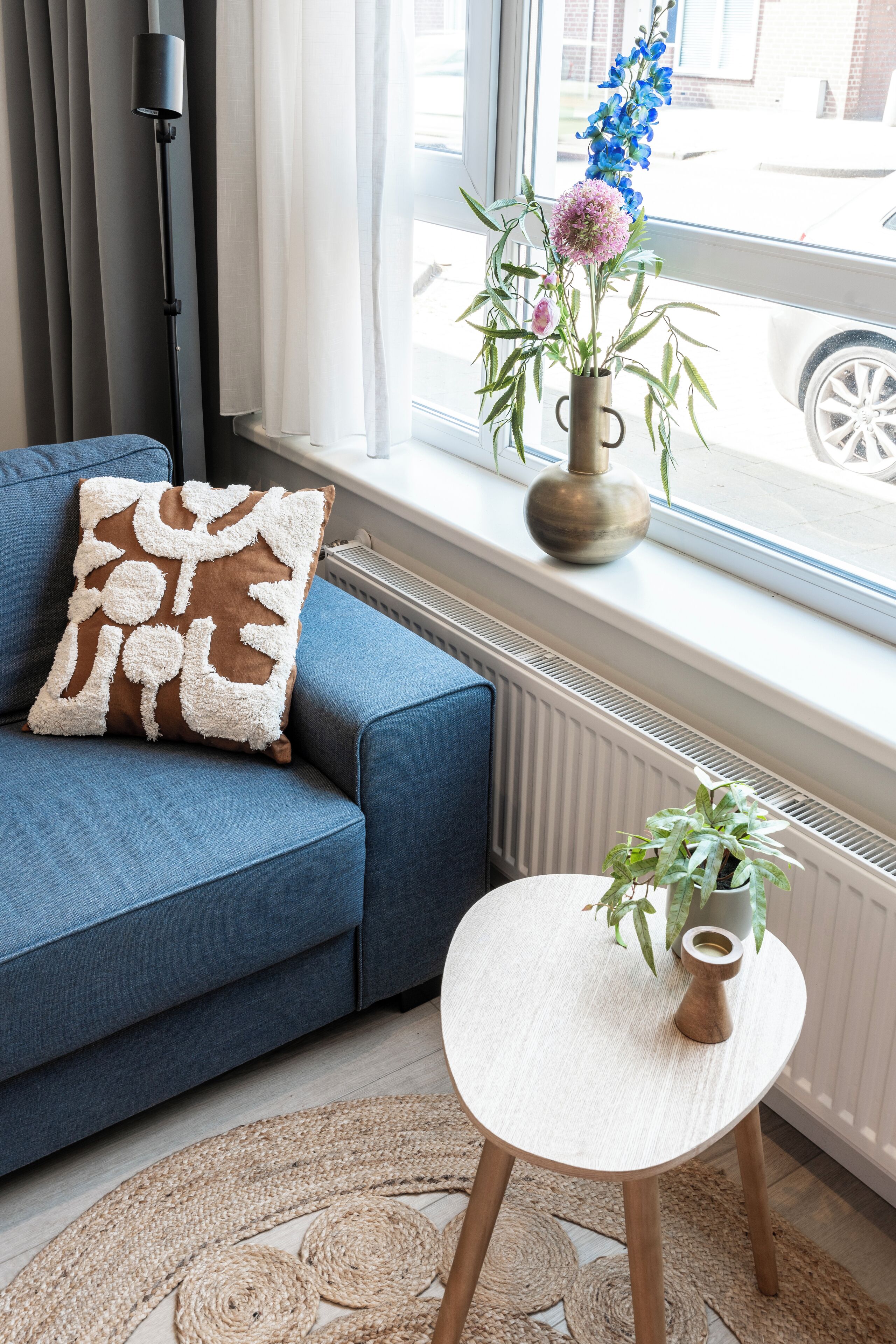 Un sofá azul con un cojín decorativo, una mesita redonda con una planta, y un florero con flores en un alféizar.