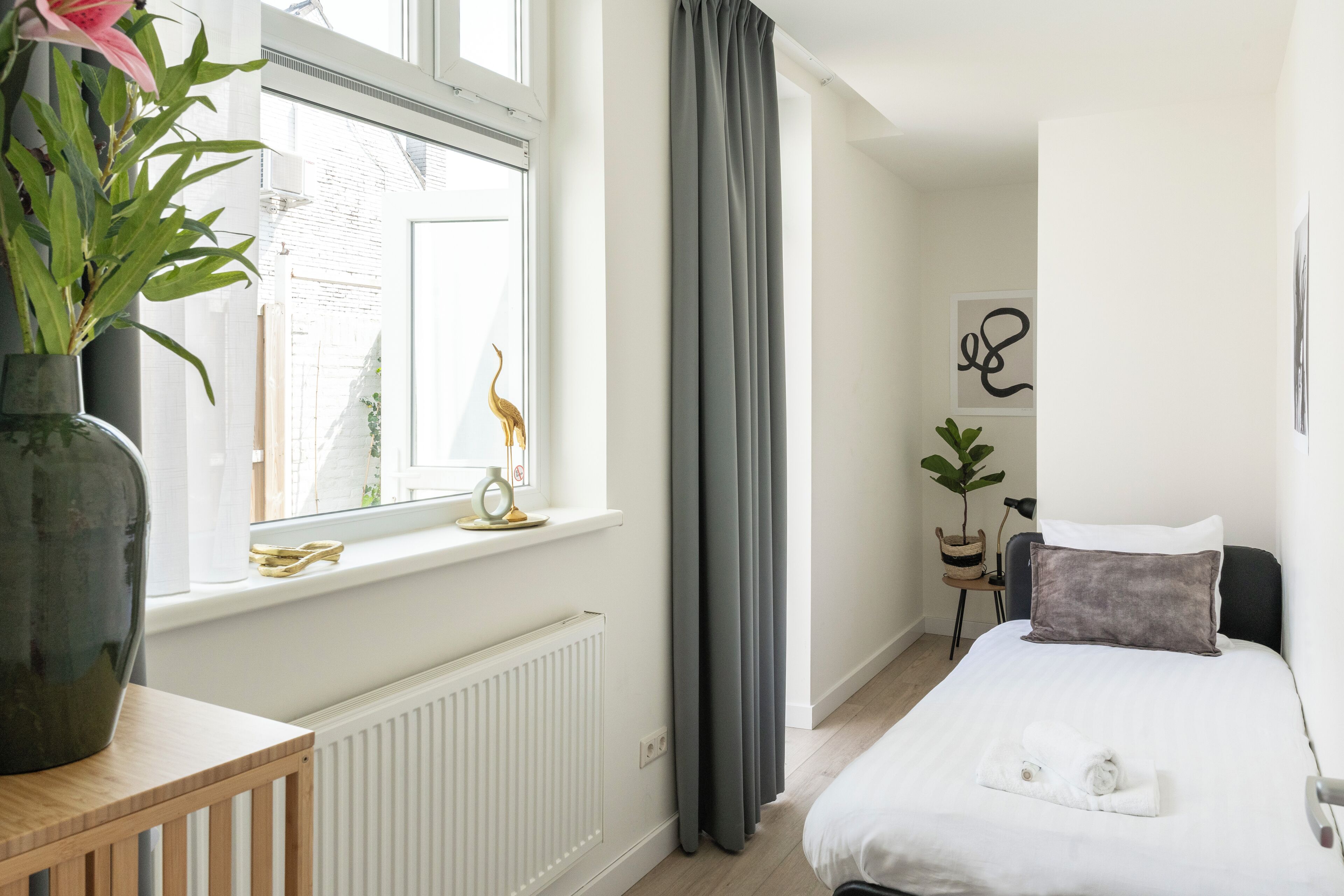 A hotel room with a bed, a window with curtains, a radiator, a plant, and a vase with flowers.
