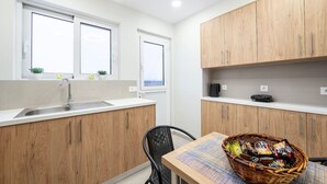 A compact kitchen with wooden cabinets, a stainless steel sink, two windows, and a dining area with a chair and a basket of snacks.