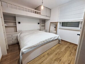 Ein Stockbett mit weißem Kopfteil, ein Nachttisch, ein Spiegel und ein Fenster mit Jalousien.