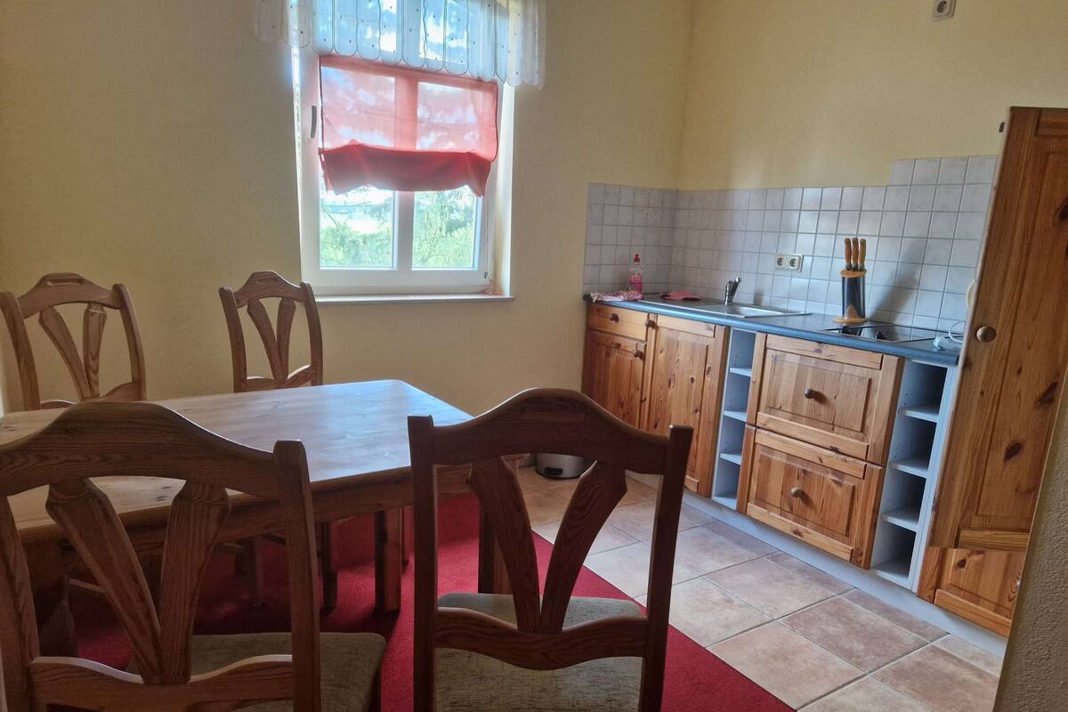 Un coin repas avec une table et des chaises en bois, une fenêtre agrémentée d’un lambrequin rouge, et une cuisine avec des meubles en bois et un dosseret carrelé.