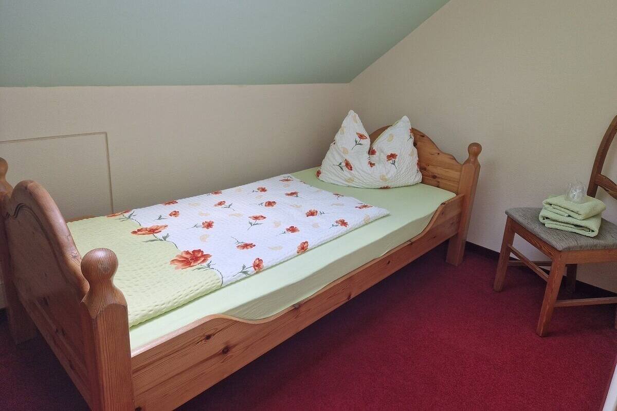Un simple lit en bois, recouvert d’une couvre-lit à motifs floraux et agrémenté d’oreillers, une chaise en bois et un sol recouvert d’une moquette rouge, le tout dans une pièce dont le plafond est en pente.