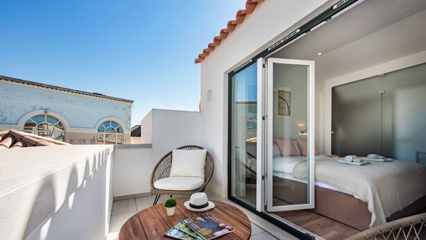A balcony with a wooden table, a wicker chair, a bed with a glass door, and a view of a building with blue walls and arches. - Faro Downtown - F in Faro (Faro)
