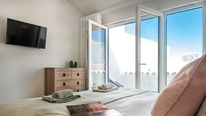 A bedroom with a bed, a flat-screen TV, a wooden dresser, a window with a view, and a magazine on the bed. - Faro Downtown - F in Faro (Faro)