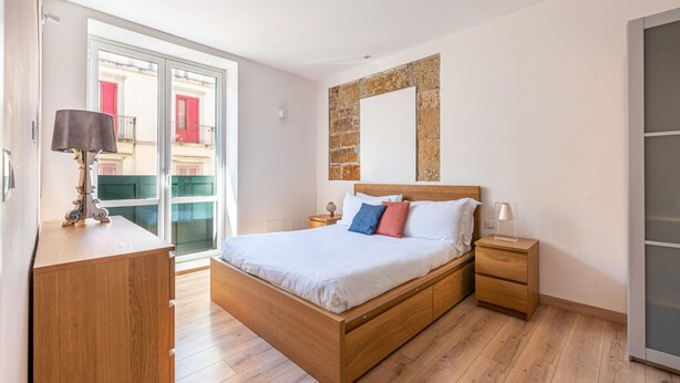 Ein Schlafzimmer mit einem Holzbett, einem Nachttisch, einer Lampe, einem Kleiderschrank und Blick auf ein Gebäude durch eine Glastür.
