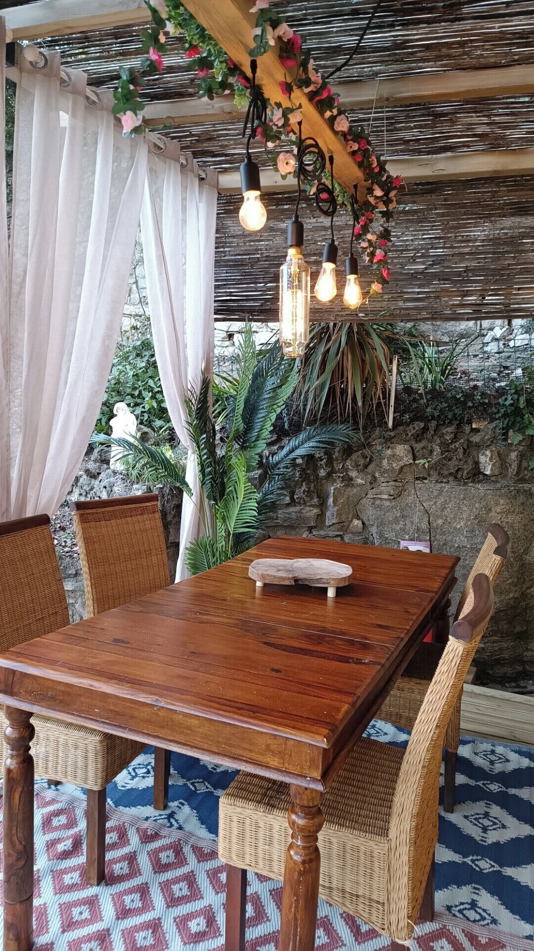 A wooden table with wicker chairs, a stone wall, and hanging lights.