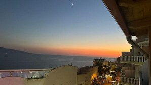 A sunset view from a balcony overlooking a coastal street with buildings and a distant mountain. - Hotel Edina by Hp (Sarande)