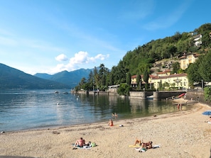 Une plage au bord d’un lac, où des gens se prélassent au soleil, avec une rive sablonneuse et des bâtiments en arrière-plan.