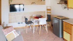 Een ruimtelijke woonkamer met een bank, eettafel, stoelen, een televisie boven de tafel, een koelkast en een keukenwerkblad.