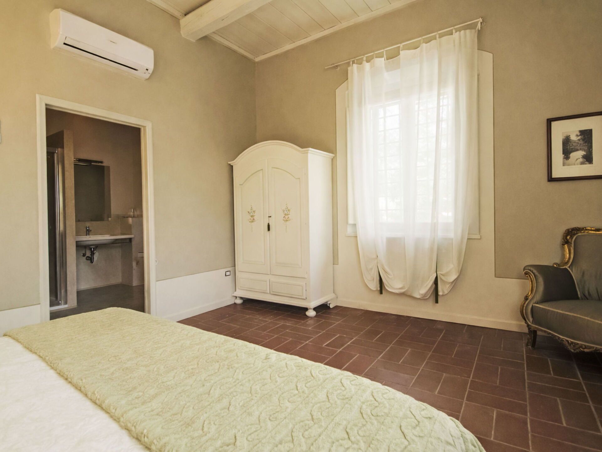Un dormitorio con cama, armario, baño y ventana con cortinas.
