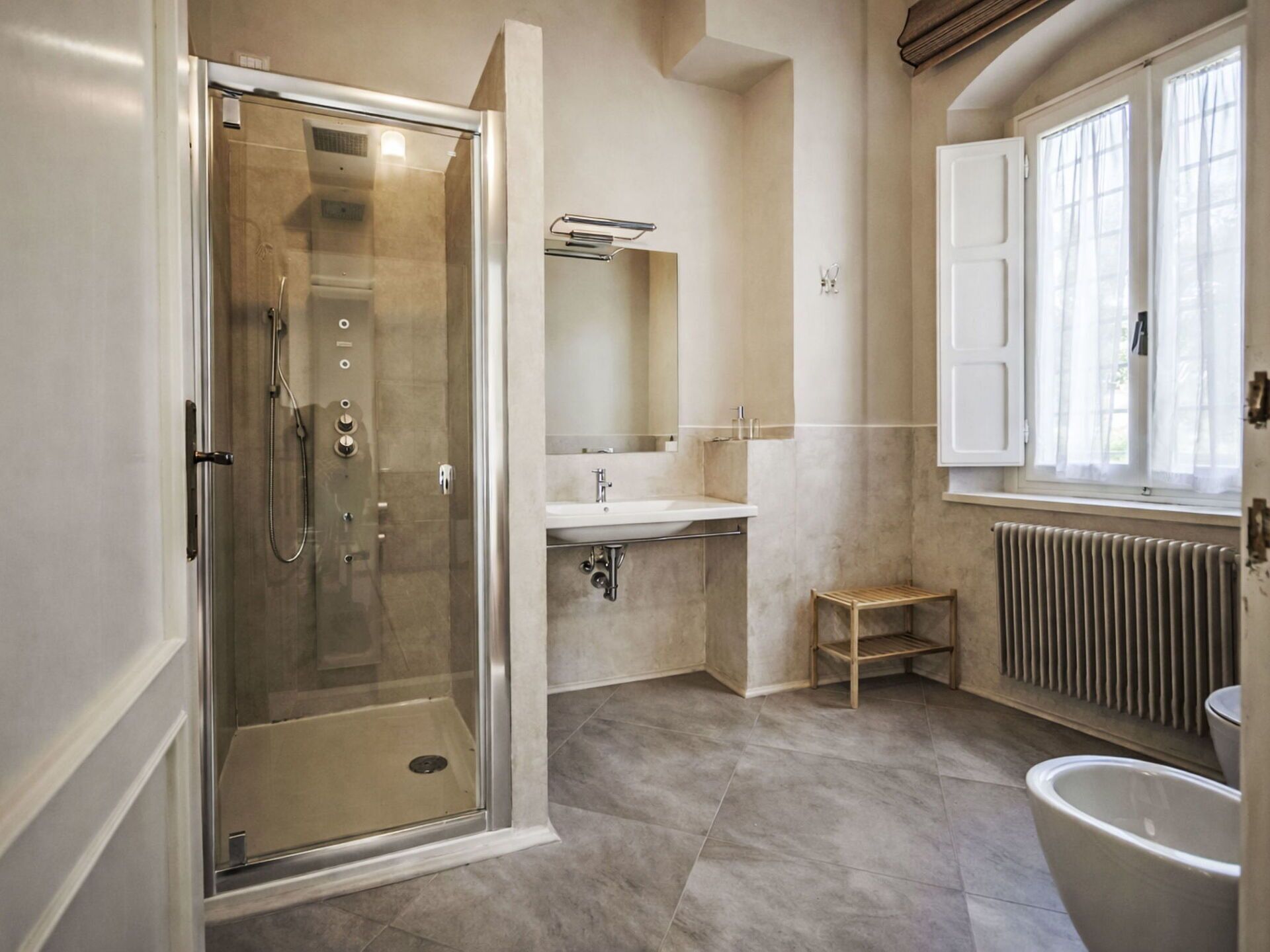 A bathroom with a glass-enclosed shower, a sink, a mirror, a radiator, and a window.