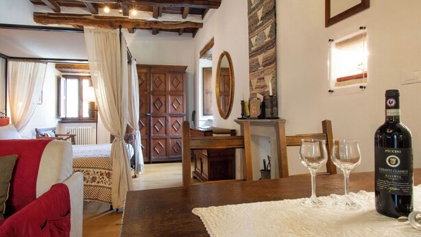 Um quarto acolhedor com teto de madeira, cama com manta vermelha, mesa com garrafa de vinho e copos, e lareira.