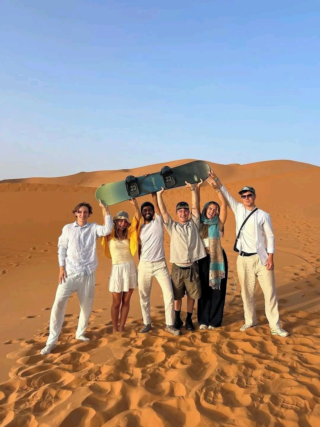 A group of people standing on a sandy beach with a snowboard held above their heads.