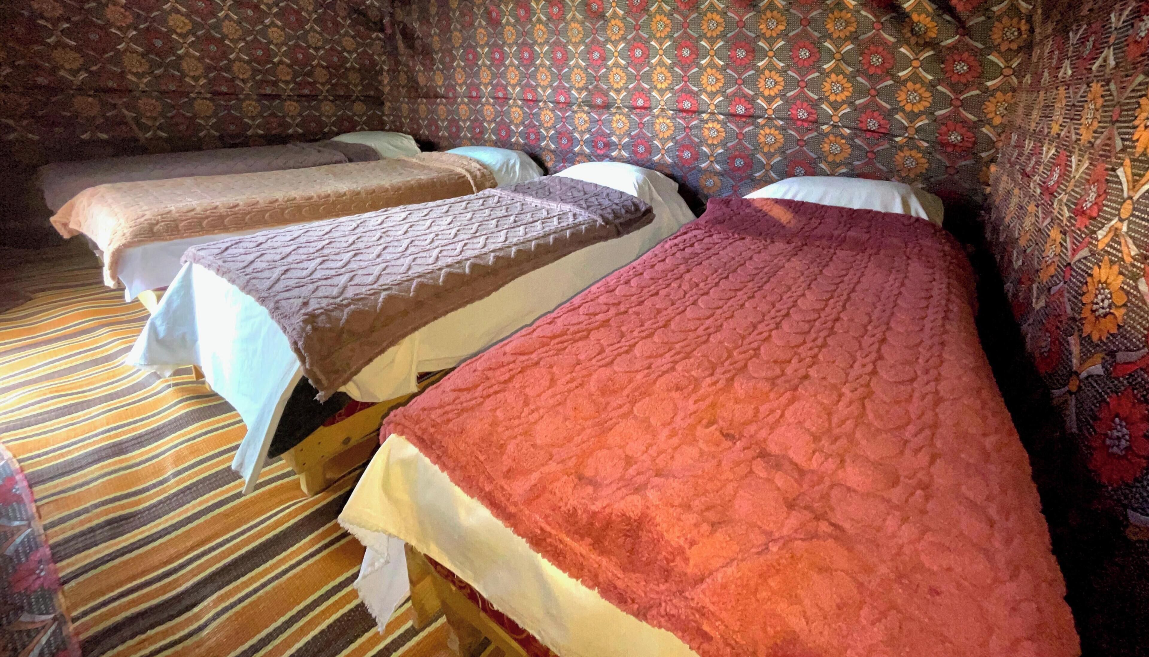 Quarto com três camas, cada uma com uma colcha de textura diferente. O quarto possui papel de parede estampado e carpete listrado.