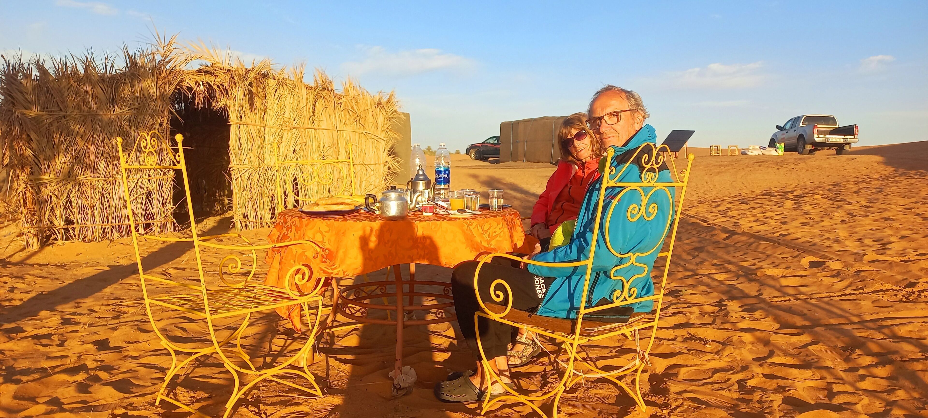 Deux personnes sont assises à une table dont le cadre est en métal, avec un panier tressé et un pot en métal, le tout dans un décor désertique.