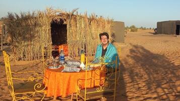 Une personne est assise à une table recouverte d’une nappe, entourée de chaises en métal, dans un paysage désertique à la végétation clairsemée, sous un ciel dégagé.