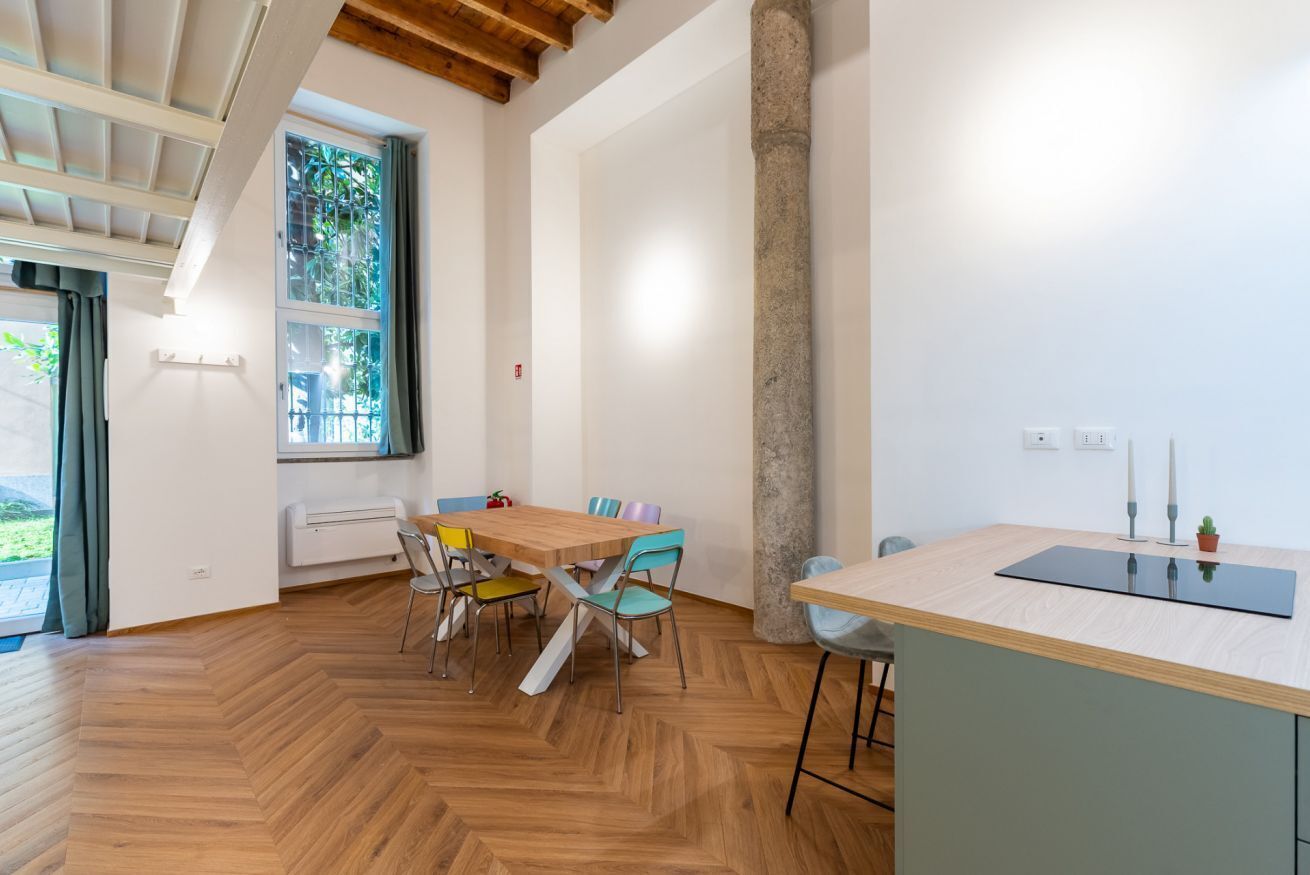 A modern interior with a wooden herringbone floor, a dining area with colorful chairs, a kitchen island, and a large window with a view.