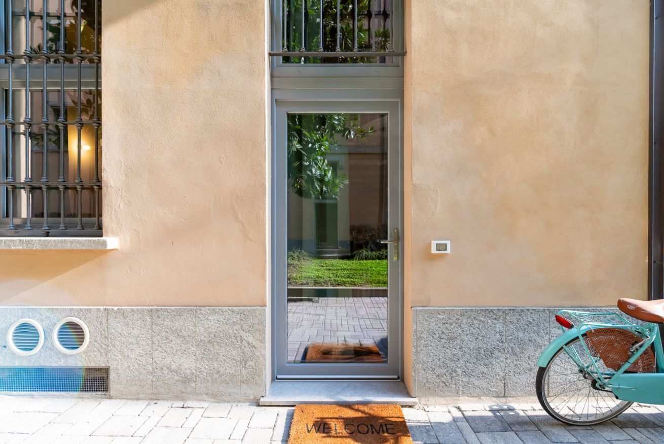 Une entrée d’immeuble avec une porte vitrée, un paillasson, un vélo et une fenêtre avec des barreaux.