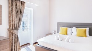 A hotel room with a bed, pillows, a nightstand, a lamp, and a window with curtains. - Duplex Sliema (Sliema)