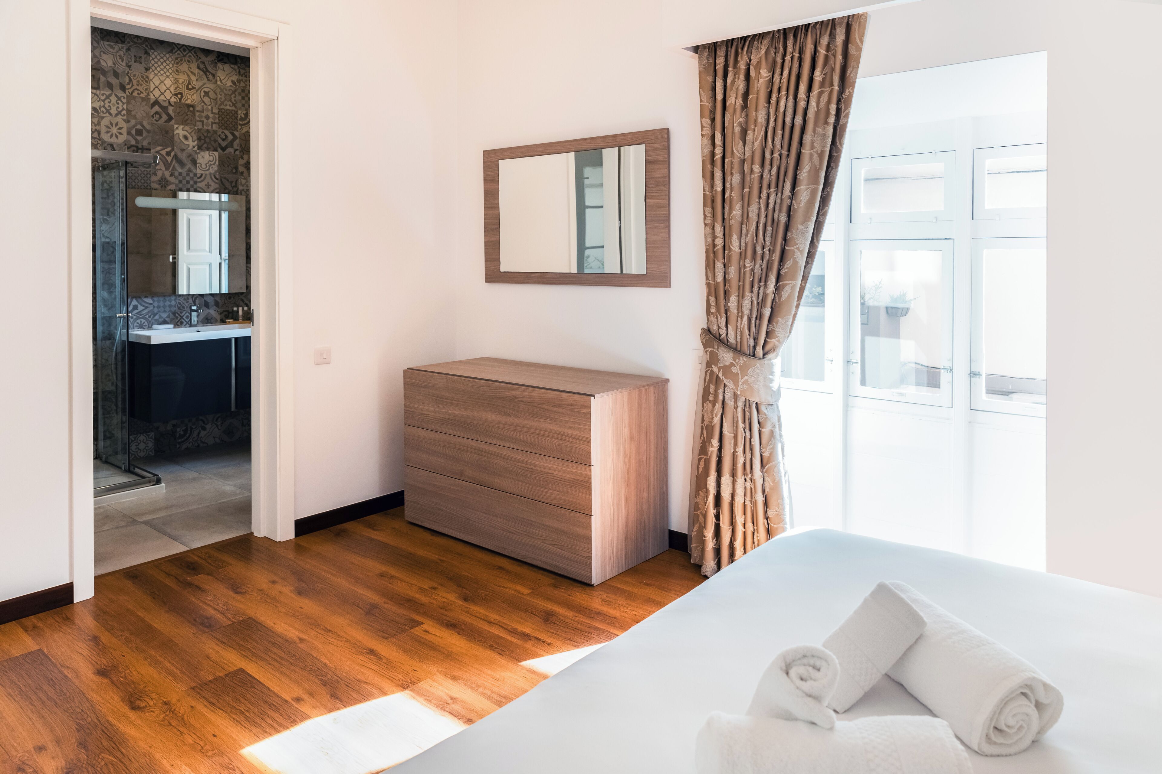 A modern hotel room with a wooden floor, a large bed, a wooden dresser, a mirror, and a view of the outside through a window.