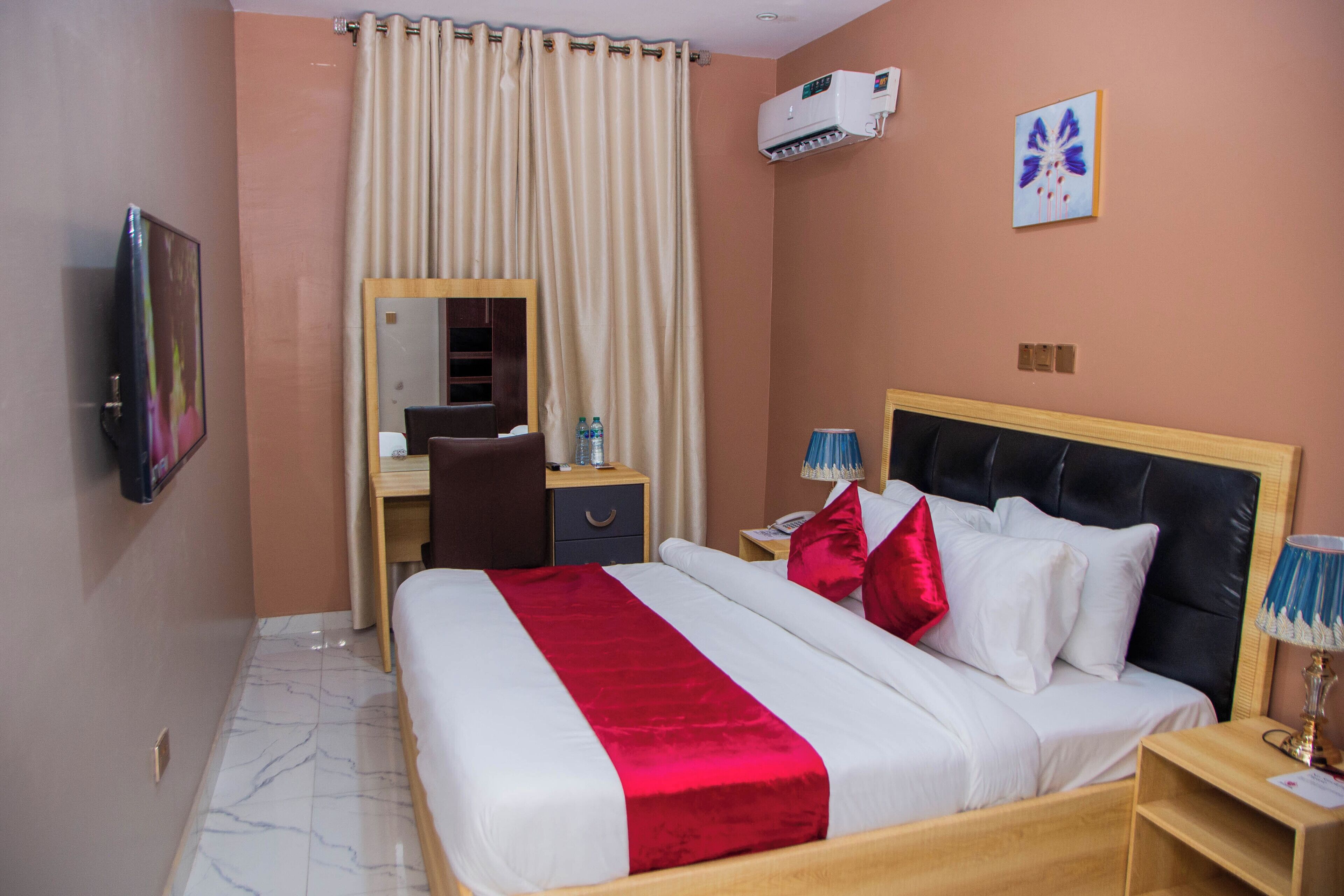 A hotel room with a bed, a desk, a chair, a mirror, a nightstand, a lamp, and an air conditioning unit.