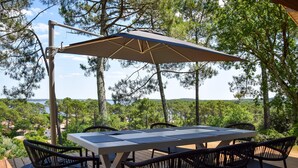 Une terrasse avec une table et des chaises, un parasol, et une vue sur des arbres et une étendue d’eau.