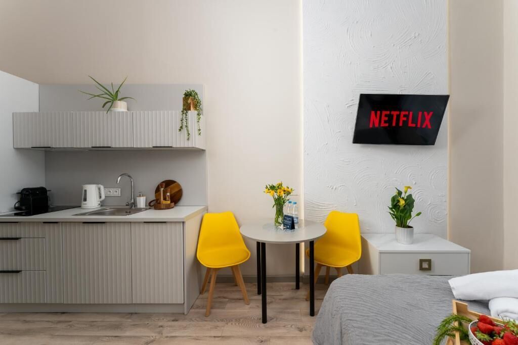 A modern hotel room with a kitchenette, a dining area with yellow chairs, a bed, and a wall-mounted TV labeled 'NETFLIX'.