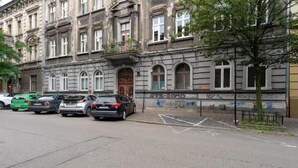 Una fila de coches aparcados a lo largo de una calle, con un edificio que presenta ventanas de arco y una puerta con marco de madera.