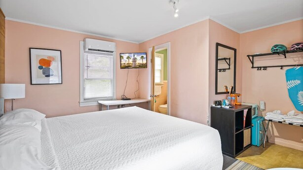 Un dormitorio con cama, escritorio, espejo, televisión y un baño visible a través de una puerta abierta.