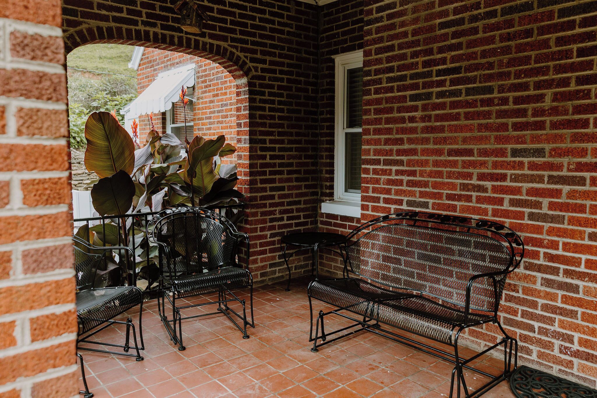 Un patio de ladrillo con sillas y un banco de metal, una ventana y un muro de ladrillo.