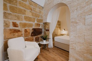 A room with a stone wall, a white sofa, a small table with a plant, and a bed with a white canopy. - Accogliente casa vicino al mare, a 2 minuti dalla spiaggia (Sicilia)