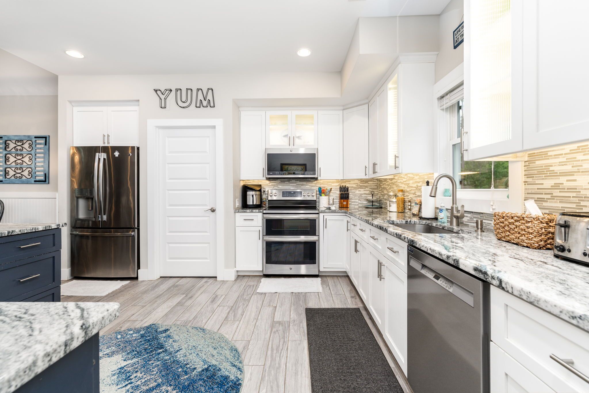 A modern kitchen with white cabinets, stainless steel appliances, and a 'YUM' sign on the wall.