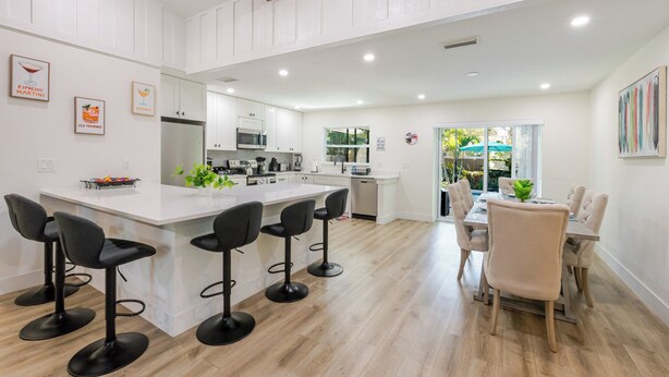 A modern kitchen with white cabinetry, a central island with bar stools, and a dining area with a table and chairs. - Summer House - Oasis pool house in the heart of Sarasota (Sarasota)