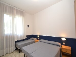 Een kamer met twee bedden, een raam met witte gordijnen, een wandlamp en een klein houten nachtkastje.