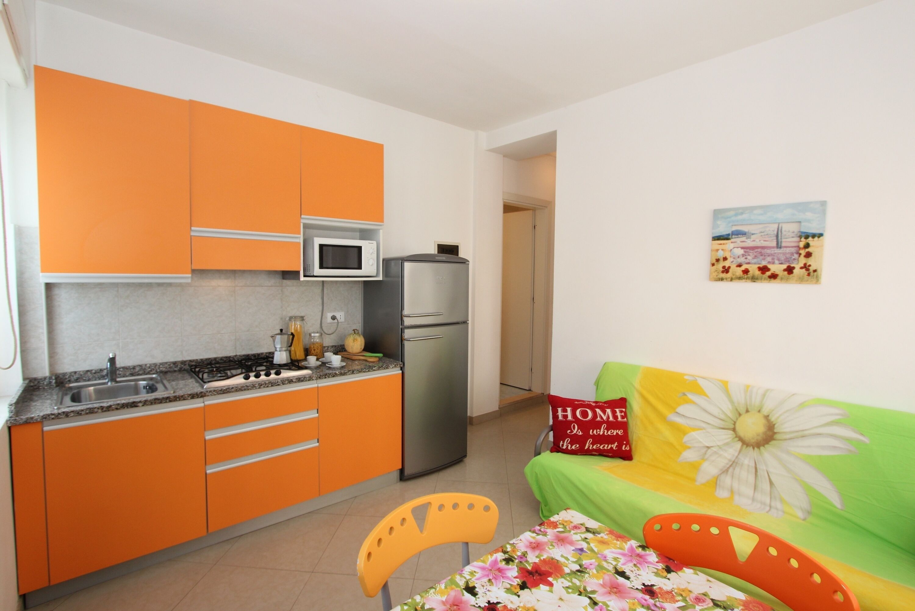 Una cocina compacta con gabinetes naranja, microondas y refrigerador. Hay un comedor con mantel floral y una silla.