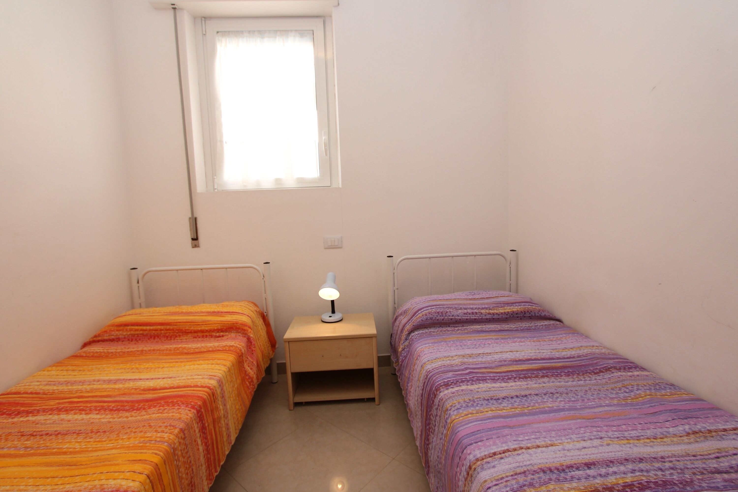 A small room with two single beds, a nightstand with a lamp, and a window with a curtain.
