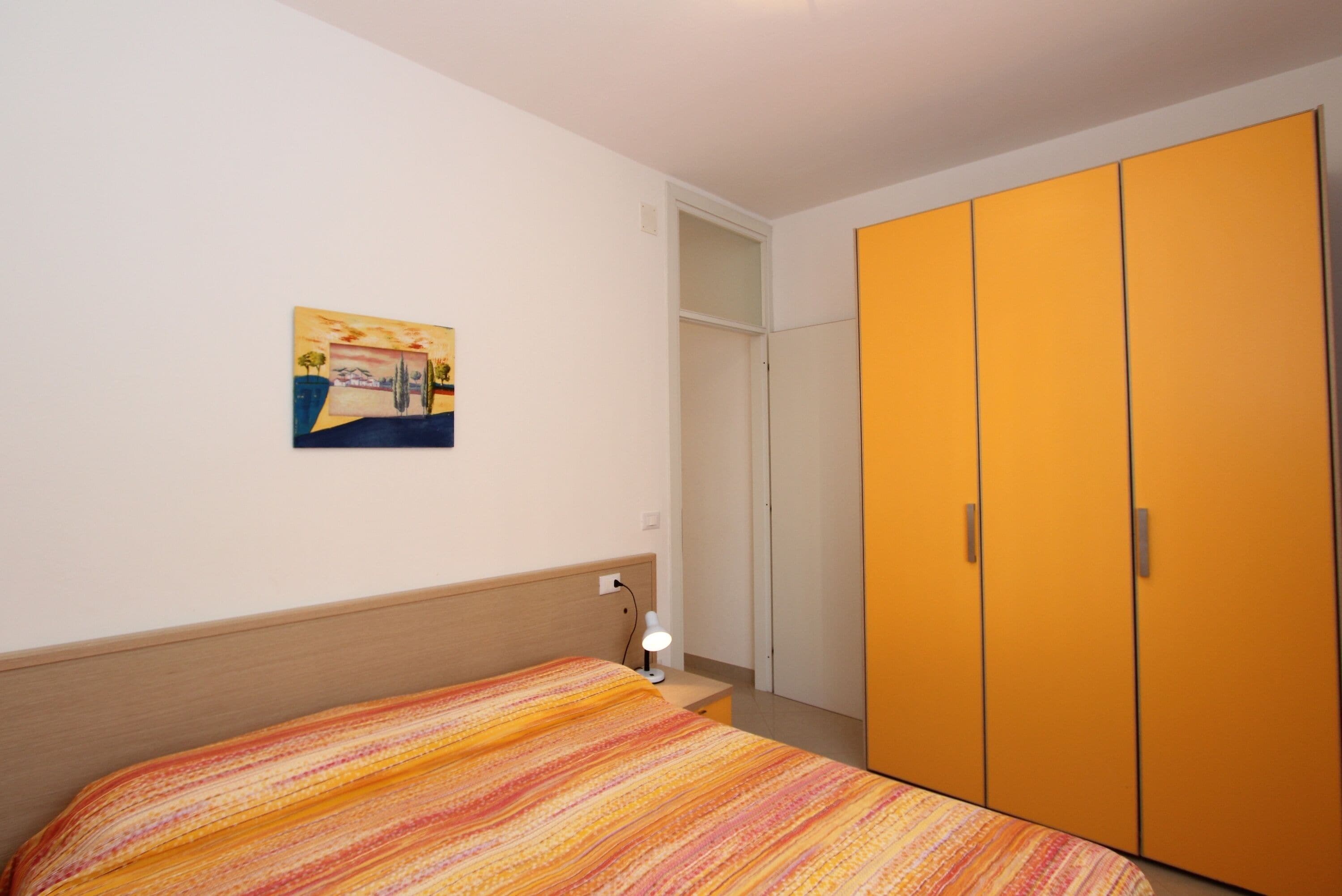 A bedroom with a bed, a nightstand, a lamp, a wardrobe, and a painting on the wall.