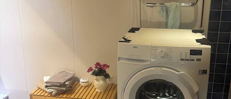 A compact laundry area with a front-loading washing machine, a drying rack, a small shelf with towels and a vase, and a towel rack on the wall.