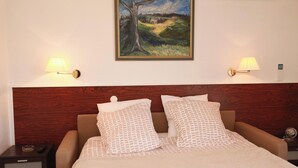 Une chambre d’hôtel avec une tête de lit en bois, deux coussins à motifs, un lit recouvert d’une courtepointe blanche, deux lampes fixées au mur et un tableau au-dessus du lit.