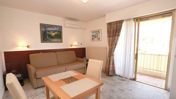 Une chambre d’hôtel avec un canapé, une table à manger, deux chaises, un tableau au mur, un climatiseur et une vue sur l’extérieur, visible à travers une porte vitrée.