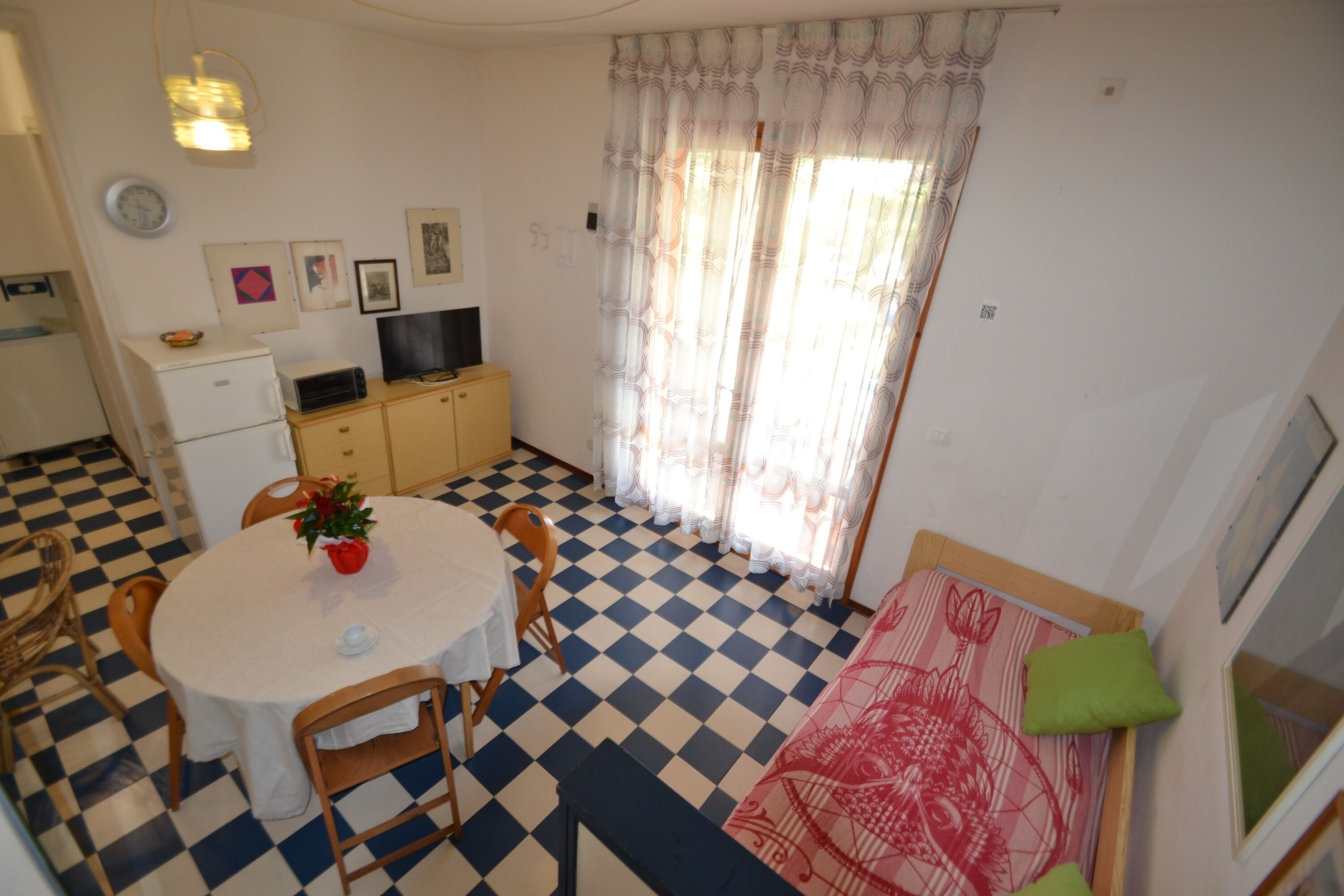Quarto com piso xadrez, mesa de jantar redonda com cadeiras, televisão, geladeira e janela com cortinas estampadas.