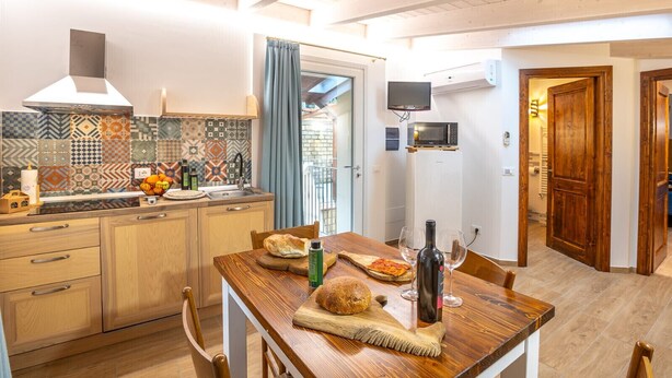 Eine Küche mit gefliestem Spritzschutz, Holzschränken, einem Esstisch mit Brot, Wein und einer Flasche sowie einem an der Wand montierten Fernseher.