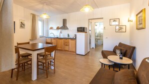 A modern kitchen with wooden cabinets, a microwave, and a stove. There is a dining area with a wooden table and chairs, and a living room with a brown sofa and a small round table with a wine bottle. - Le Vigne - Appartamento La Maremma (Bolsena)